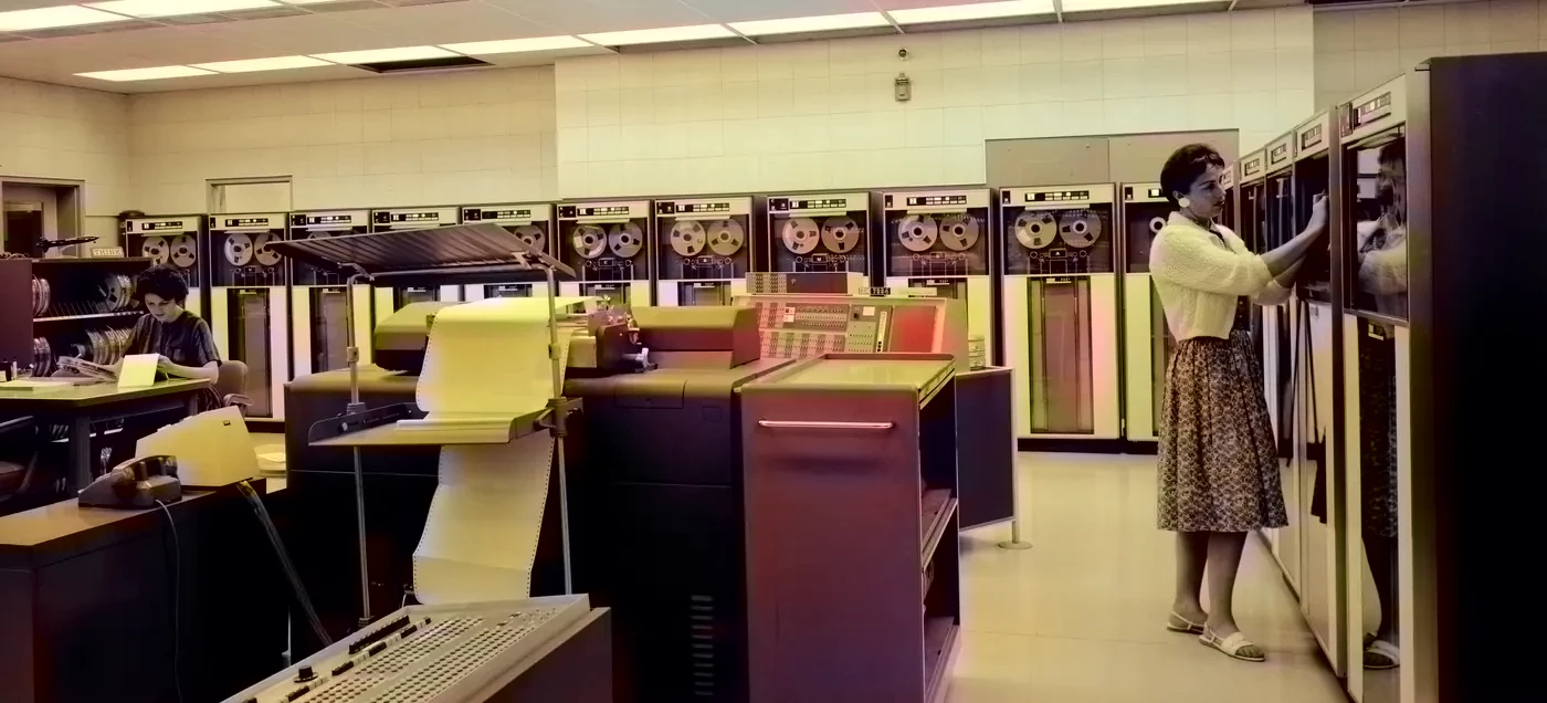 A 1963 computer room: tape drives along the wall, a woman loading a reel, a printer and an operator in the foreground.