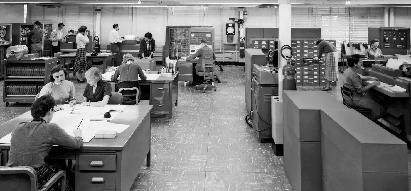 A computing room at NACA Langley in 1957: mathematicians at desks in the foreground, operators at an IBM 704 console, tape drives, and card equipment behind them.