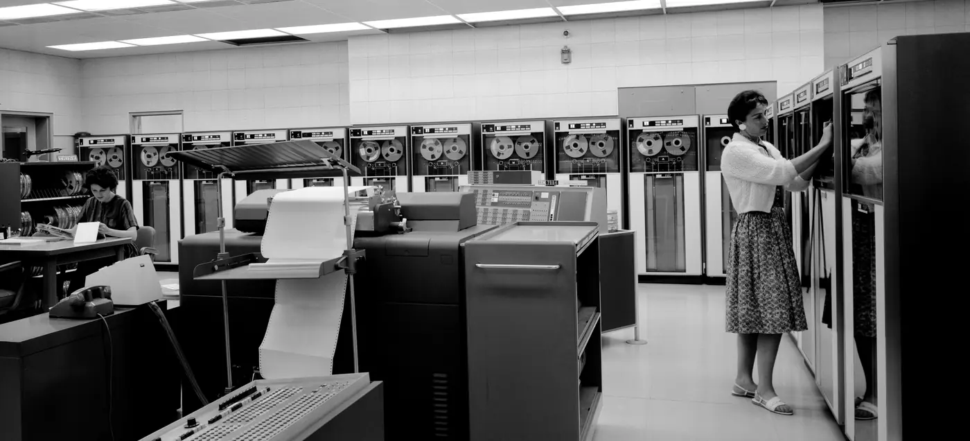 A 1963 computer room: tape drives along the wall, a woman loading a reel, a printer and an operator in the foreground.